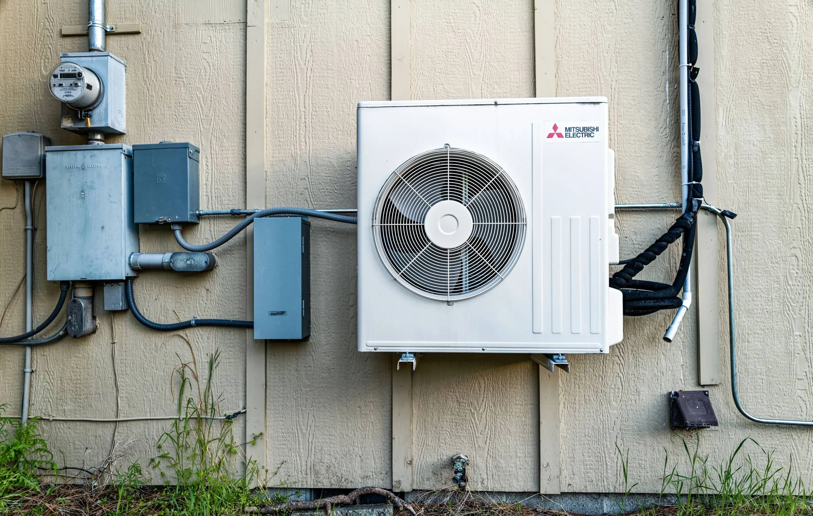 Mitsubishi Electric outdoor air conditioning unit mounted on a residential wall with electrical connections and conduit pipes. 