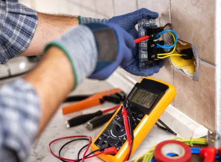 Electrician wearing gloves using a multimeter to test electrical wiring inside a wall socket.