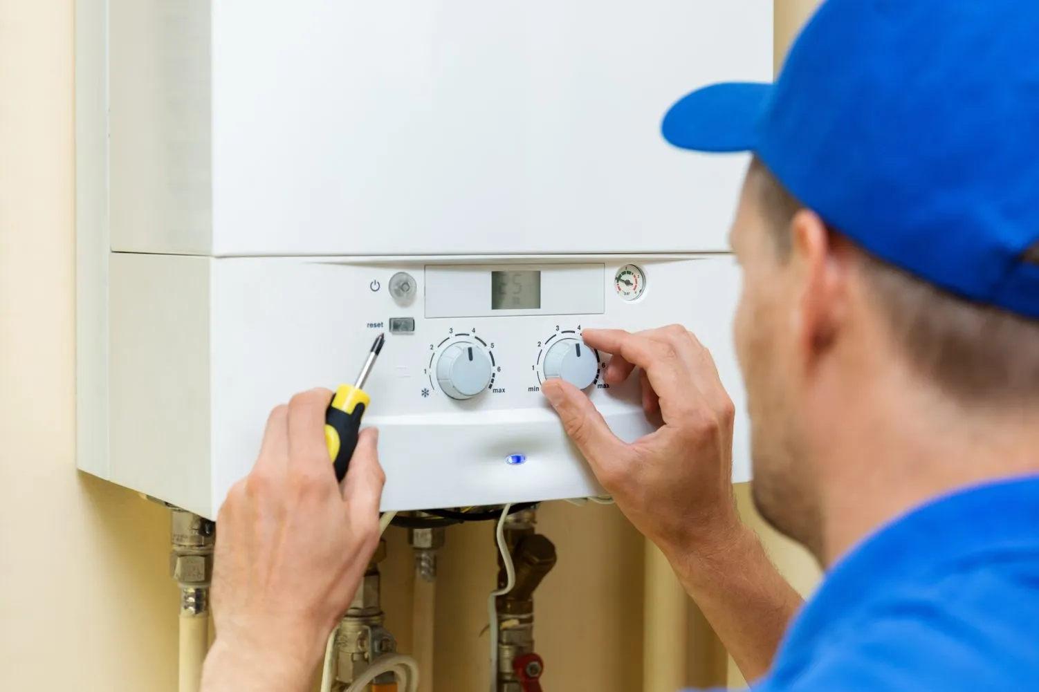 Technician in a blue uniform adjusting settings on a wall-mounted boiler with a screwdriver in hand.